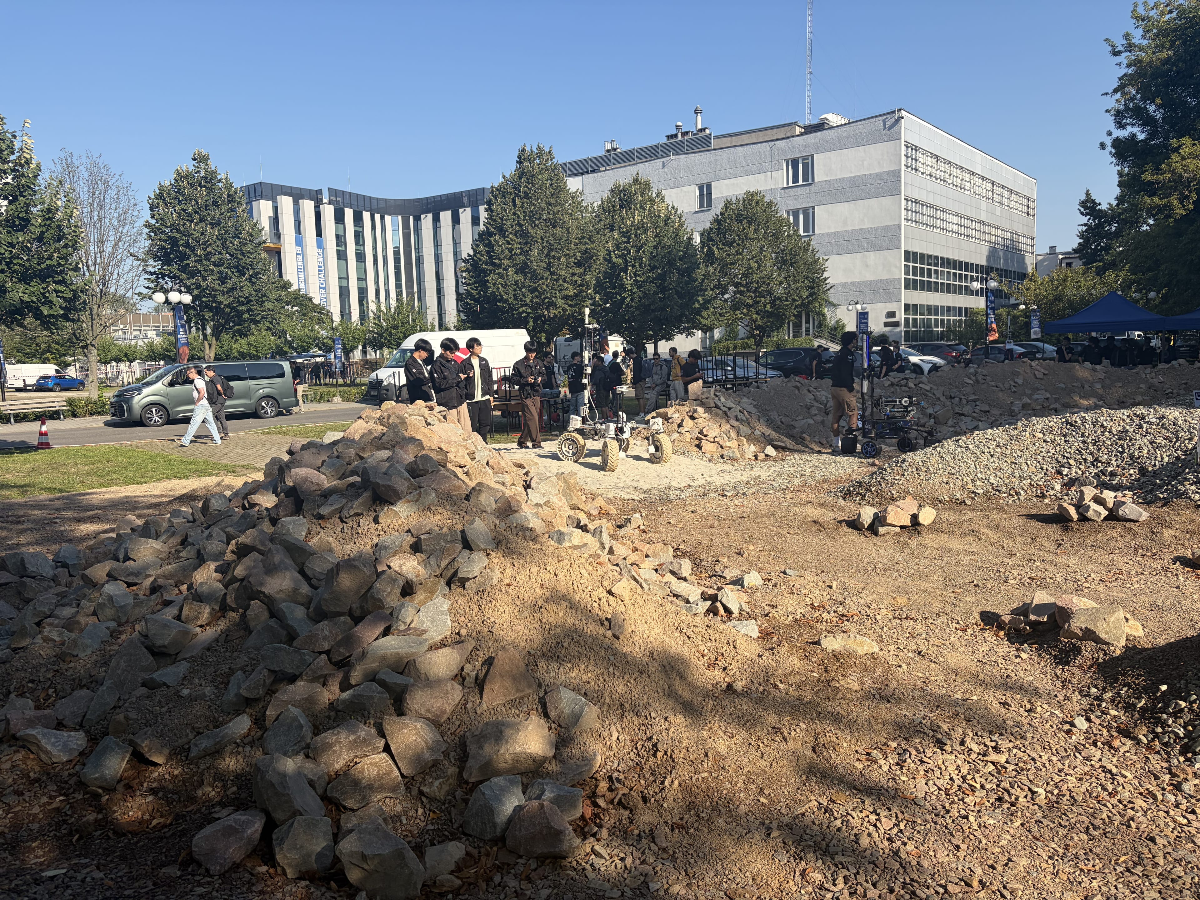 Another team runs its rover on the gravel mounds of the Mars yard with the AGH campus behind.