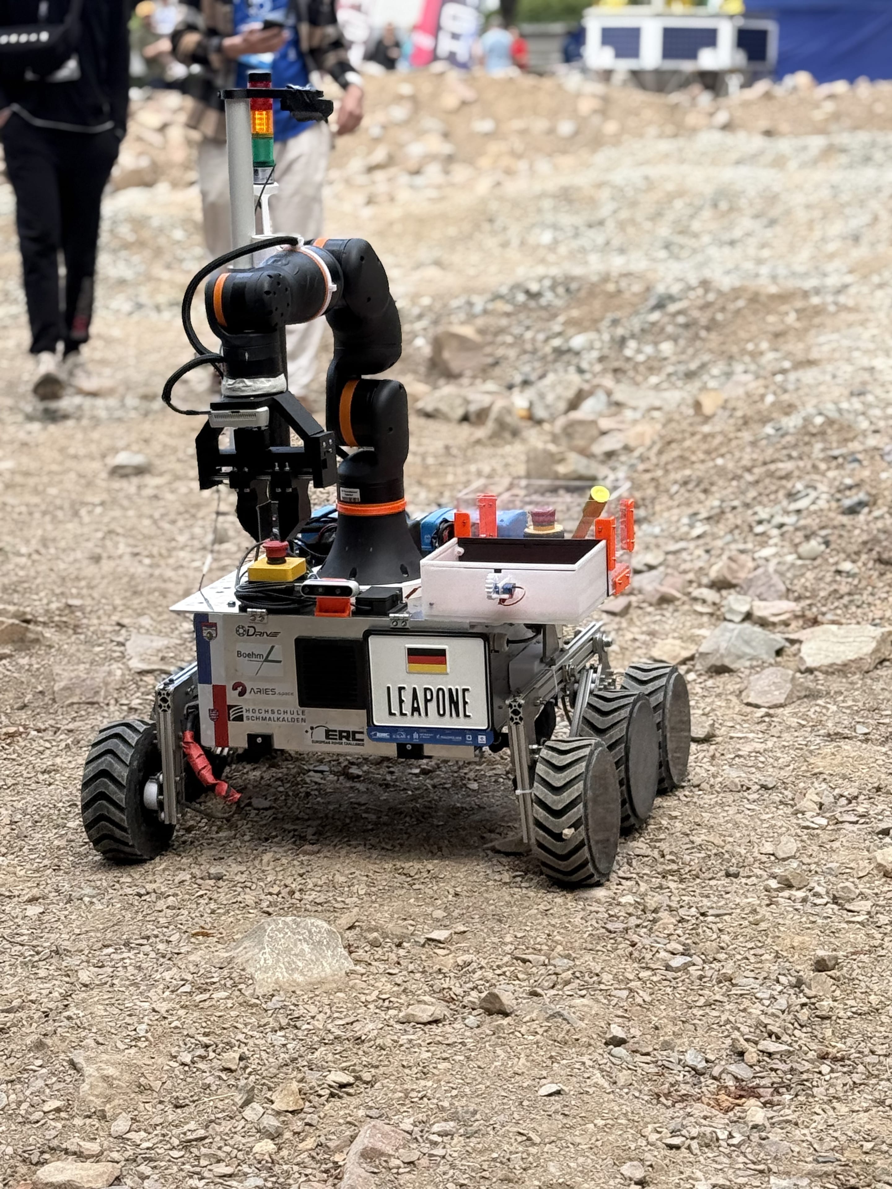 LEAP-One faces the camera on the gravel with its arm extended and a sample container on top.