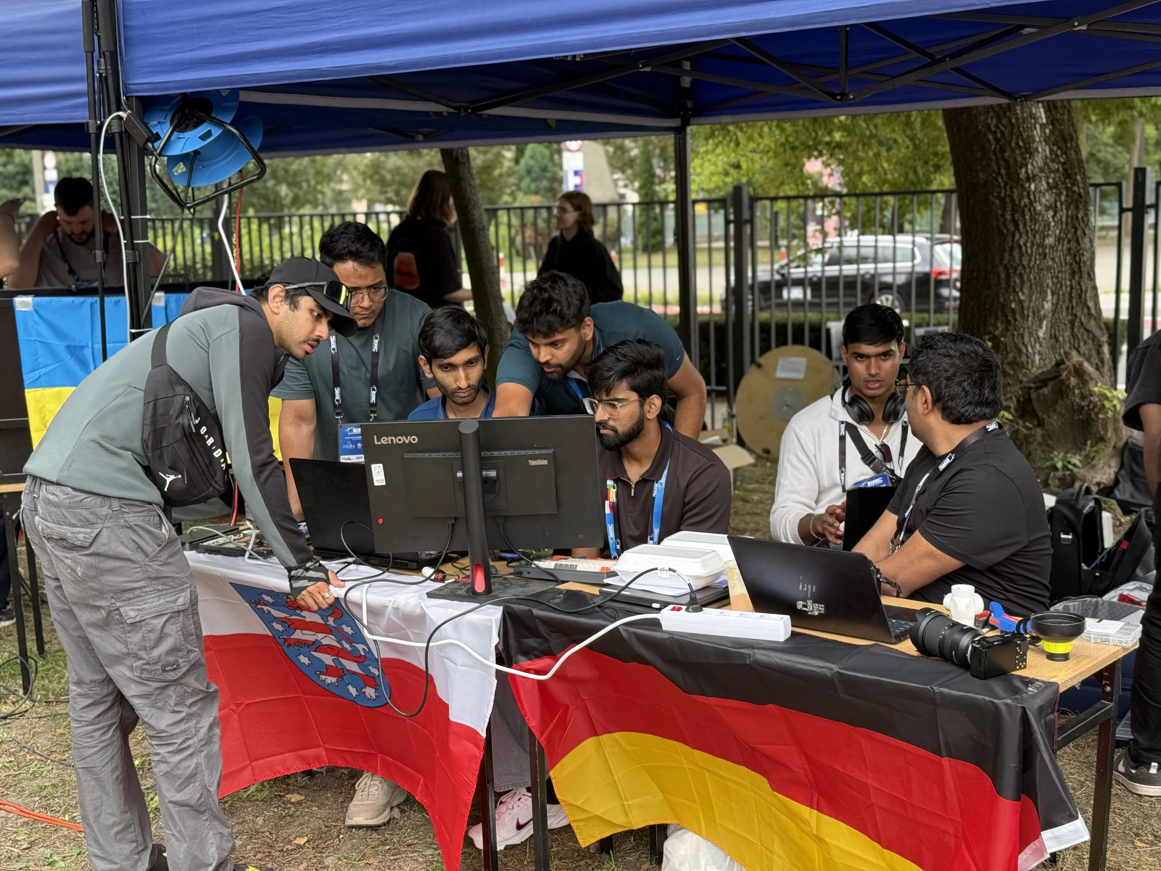 The crew huddles around the control-station monitor in the pit tent, the table draped in the Thuringian and German flags.