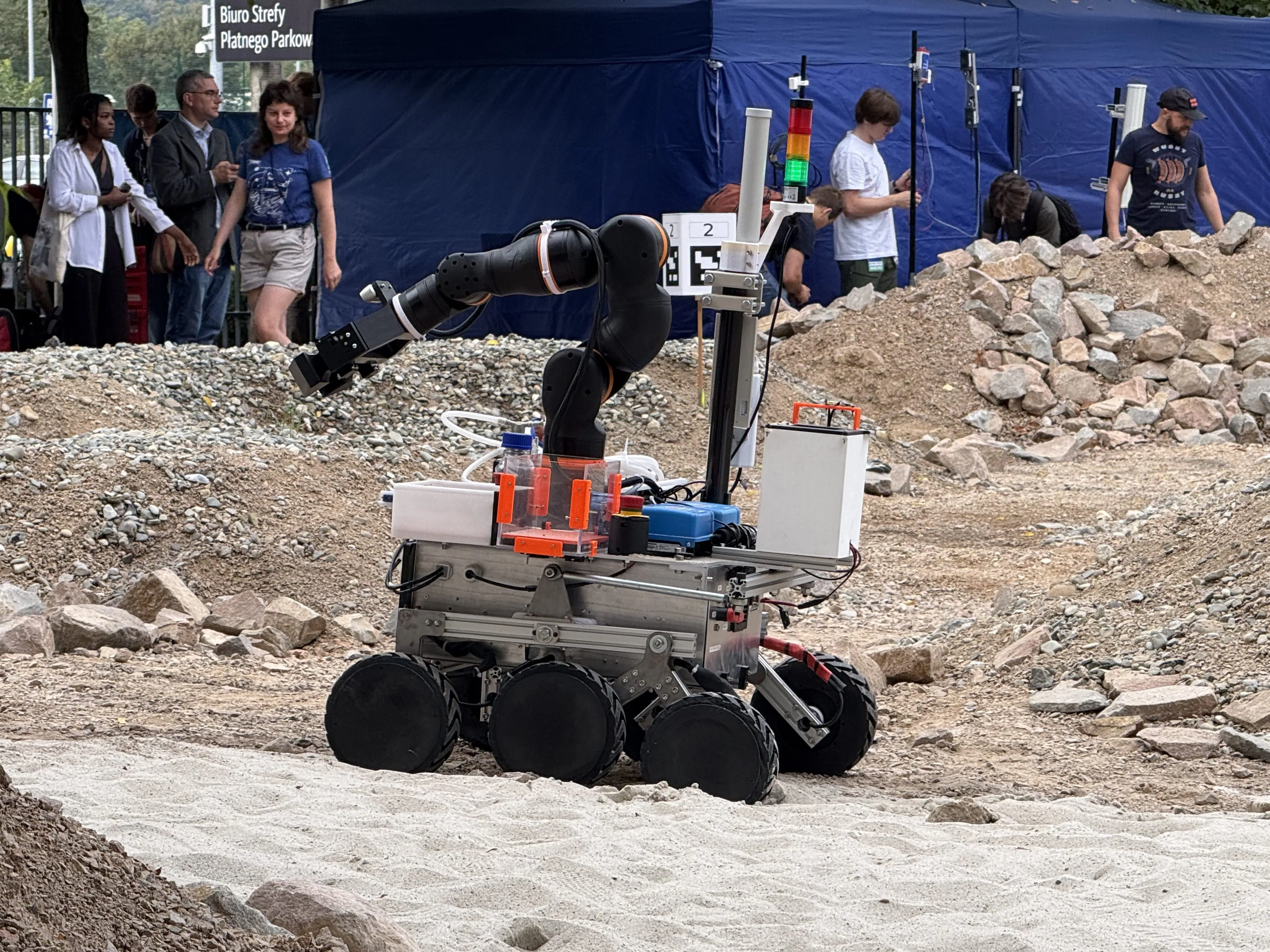 LEAP-One crosses the sand and rock of the Mars yard with its arm raised and a marker on its mast.